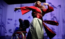 Image description: Gaelin and Maryam, ensemble members, dance together in front of a projection of black birds on a white background. Gaelin's red sash is swinging with her movements, her arm stretches out to her right and her other is folded in towards her face which is looking upwards catching the light. Behind her, Maryam, who has dark hair and wears a blue-green costume with a yellow sash, is dancing in her red wheelchair.