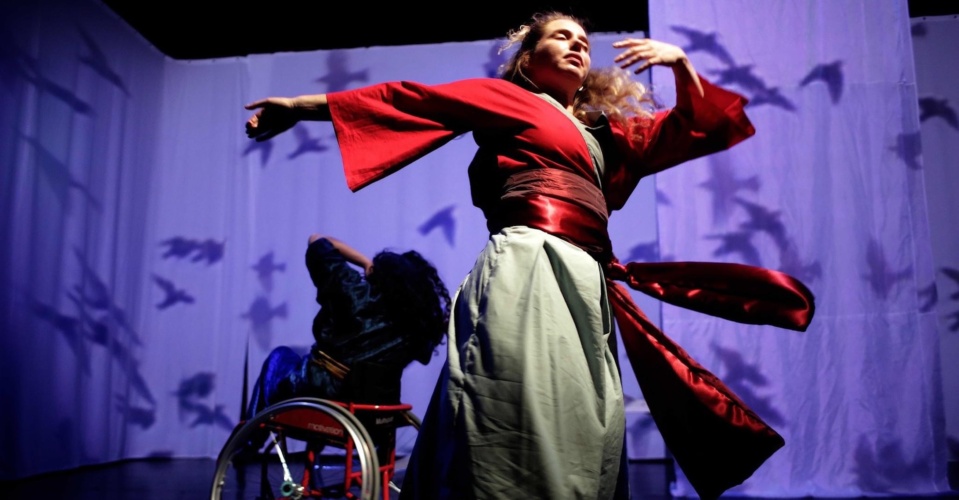 Image description: Gaelin and Maryam, ensemble members, dance together in front of a projection of black birds on a white background. Gaelin's red sash is swinging with her movements, her arm stretches out to her right and her other is folded in towards her face which is looking upwards catching the light. Behind her, Maryam, who has dark hair and wears a blue-green costume with a yellow sash, is dancing in her red wheelchair.